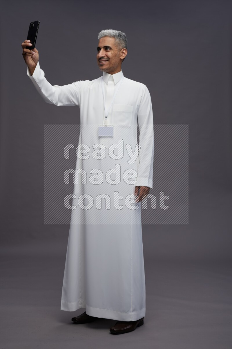 Saudi man wearing thob with neck strap employee badge standing taking selife on gray background