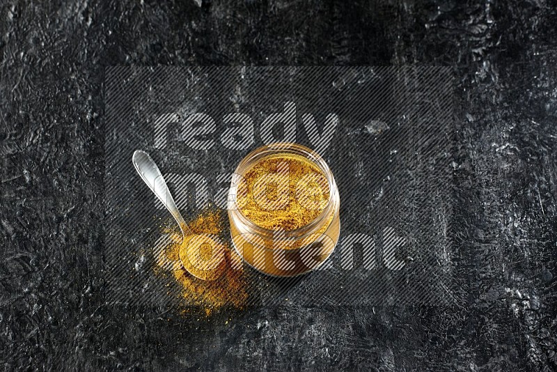 A glass jar and a metal spoon full of turmeric powder on a textured black flooring