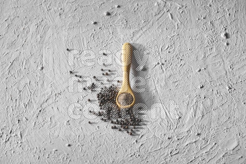 A wooden spoon full of black pepper powder and black pepper beads beside it on a textured white flooring