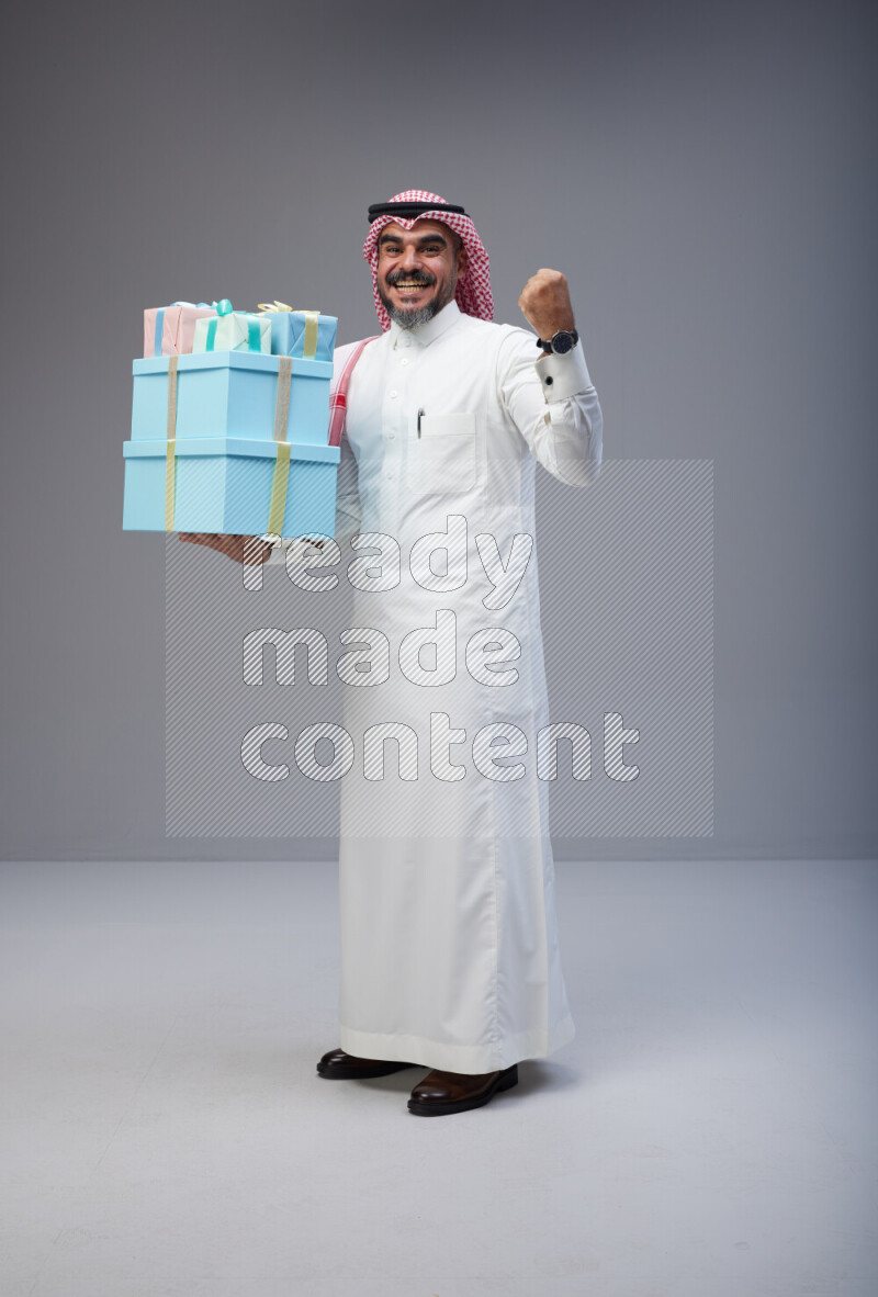 Saudi man Wearing Thob and red Shomag standing holding gift box on Gray background