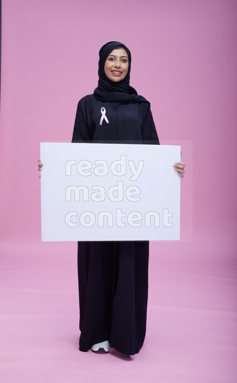 Saudi woman wearing pink ribbon on Abaya standing holding board on pink background