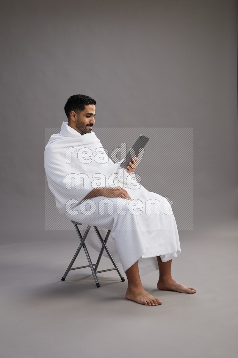 A man wearing Ehram sitting on chair holding a tablet on gray background