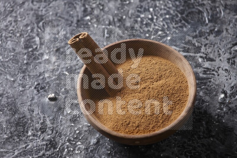 Wooden bowl full of cinnamon powder and a cinnamon stick on a textured black background