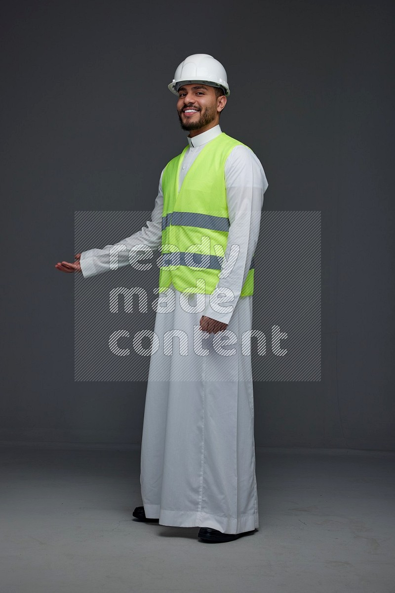 A Saudi man wearing Thobe with a yellow safety vest and white helmet standing and pointing different angles eye level on a gray background