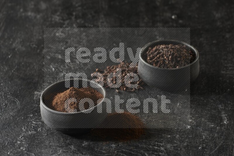 2 Black pottery bowls full of cloves and the other full of cloves powder on textured black flooring