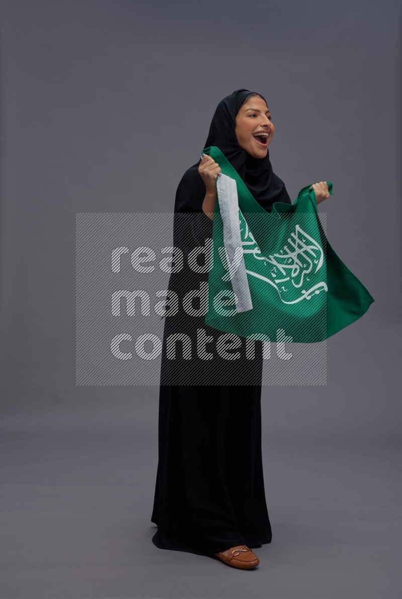 Saudi woman wearing Abaya standing holding Saudi flag on gray background