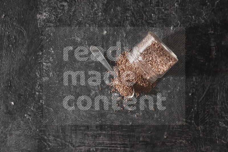 وعاء زجاجي مقلوب ممتلئ بحبوب بذر الكتان والحبوب متناثرة منه مع ملعقة معدنية ممتلئة علي خلفية سوداء