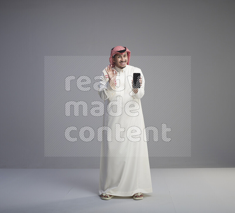 A Saudi man standing wearing thob and red shomag showing phone to camera on gray background