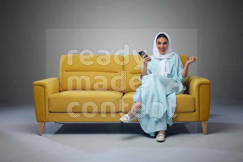 A Saudi woman wearing a light blue Abaya and white head scarf sitting on a yellow sofa and using her phone eye level on a grey background