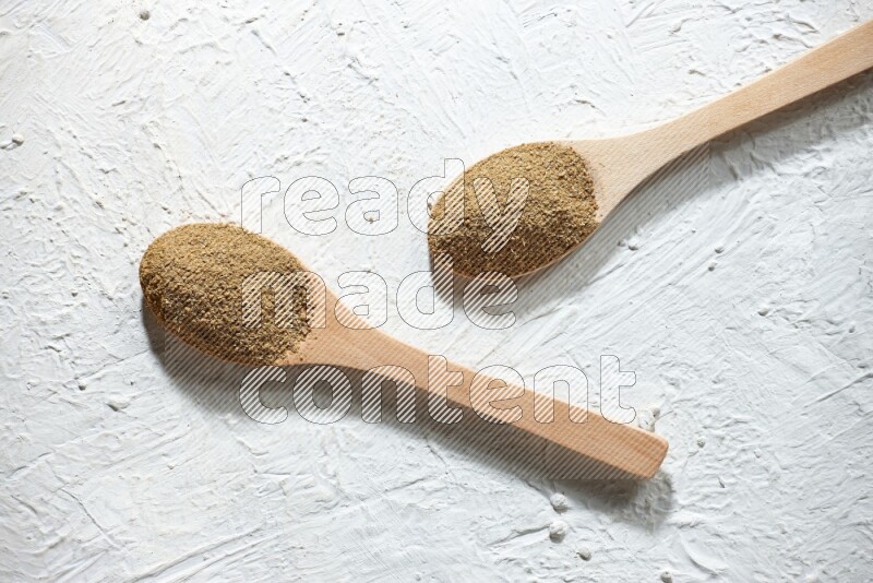 2 Wooden spoons full of cumin powder on a textured white flooring