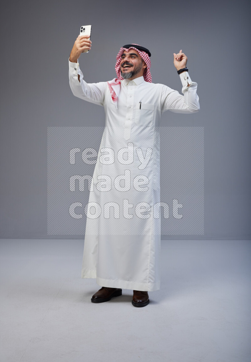 Saudi man Wearing Thob and red Shomag standing taking selfie on Gray background