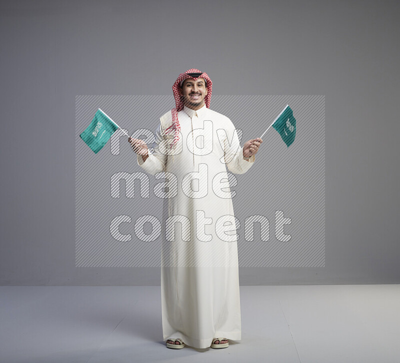 رجل سعودي يرتدي ثوب ابيض وشماغ احمر يمسك بعلمين صغيرين للمملكة العربية السعودية