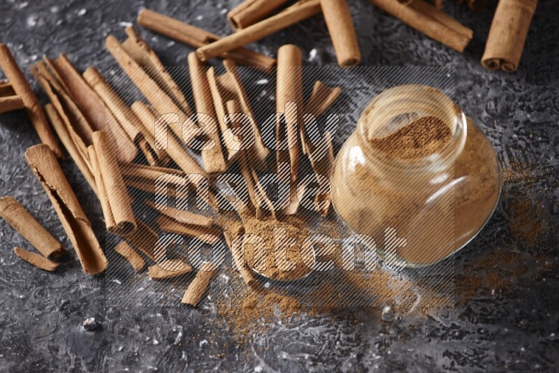 Herbal glass jar and a metal spoon full of cinnamon powder surrounded by cinnamon sticks on textured black background