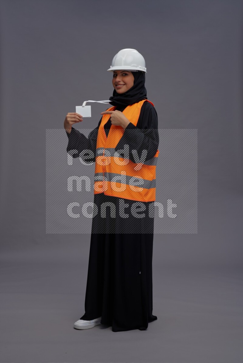 Saudi woman wearing Abaya with engineer vest with neck strap employee badge standing interacting with the camera on gray background