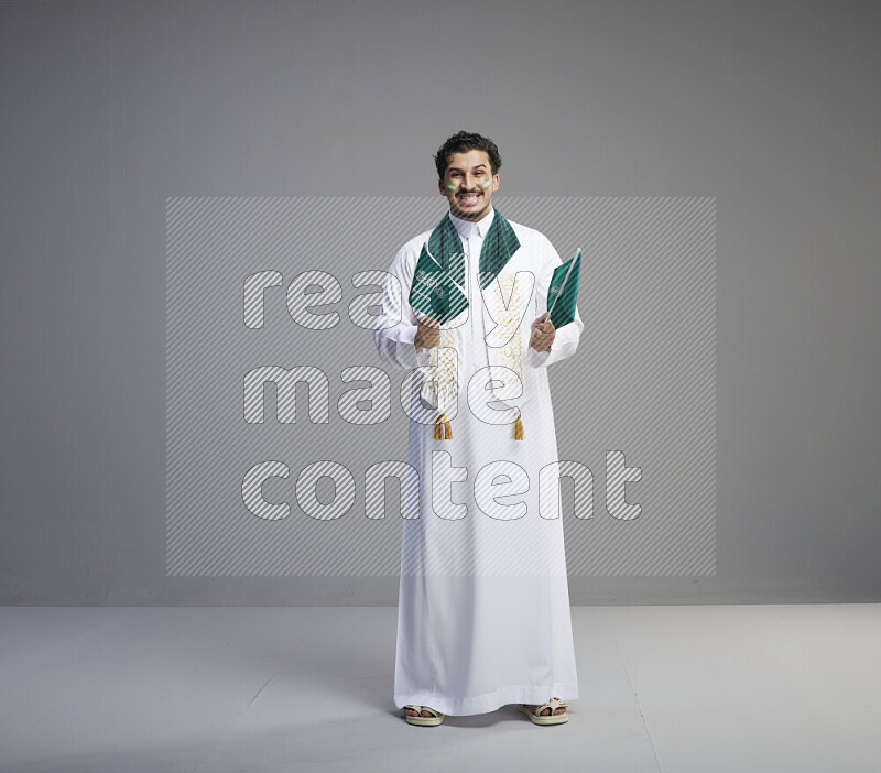 A Saudi man standing wearing thob and saudi flag scarf with face painting holding small Saudi flag on gray background