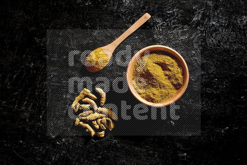 A wooden bowl and a wooden spoon full of turmeric powder with dried turmeric fingers on textured black flooring