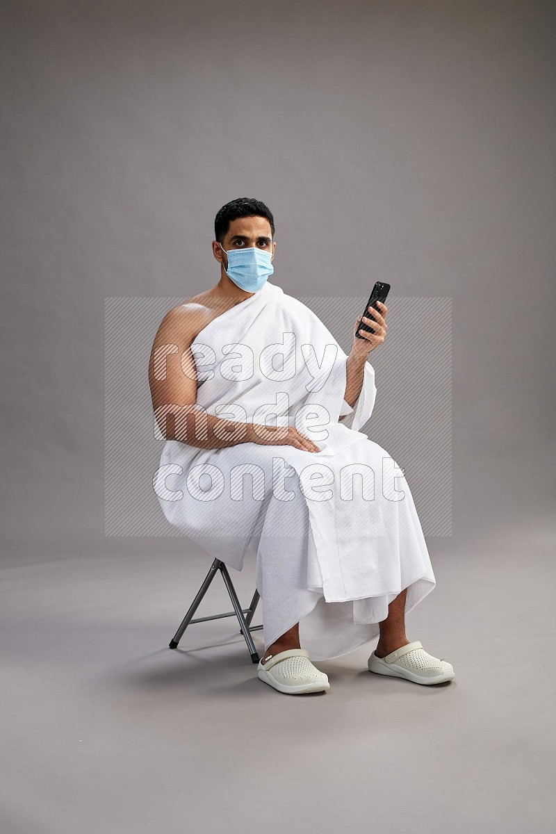 A man wearing Ehram with face mask sitting on chair texting on phone on gray background