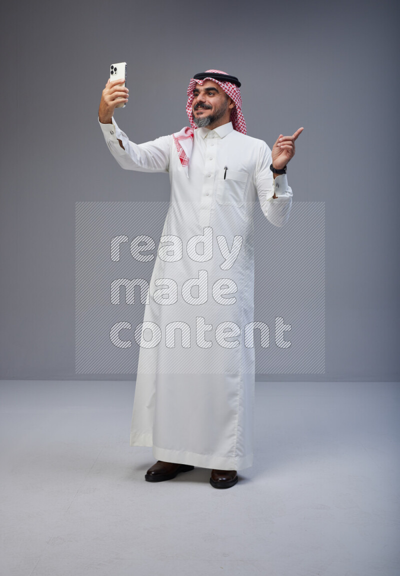 Saudi man Wearing Thob and red Shomag standing taking selfie on Gray background