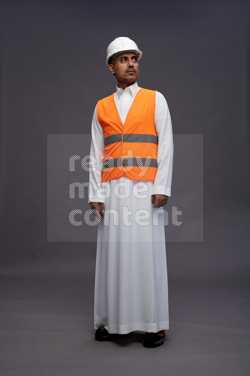 Saudi man wearing thob with engineer vest standing interacting with the camera on gray background