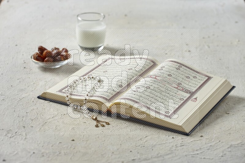Quran with dates, prayer beads and different drinks all placed on textured white background
