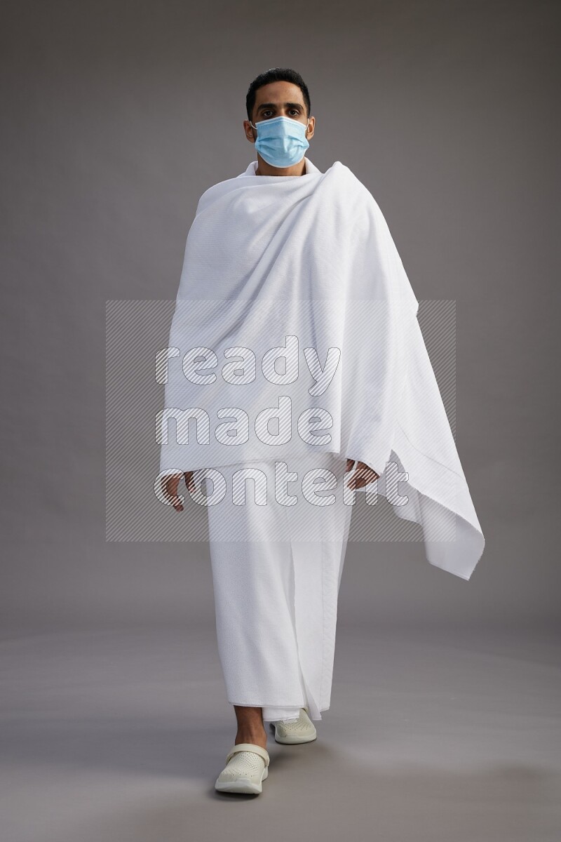 A man wearing Ehram with face mask Standing Interacting with the camera on gray background