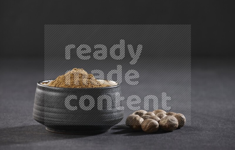 وعاء فخاري أسود ممتلئ ببودرة جوزة الطيب مع بعض حبوب جوزة الطيب بجانبه على خلفية سوداء