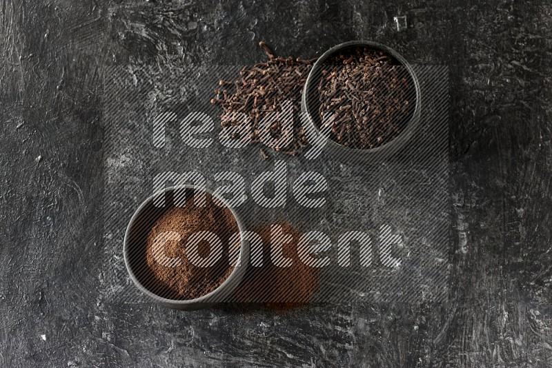 2 Black pottery bowls full of cloves and the other full of cloves powder on textured black flooring