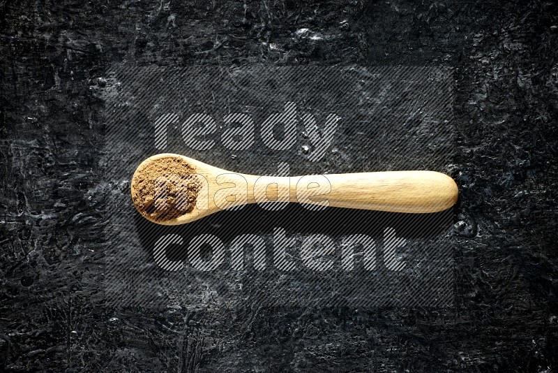 A wooden spoon full of allspice powder on a textured black flooring