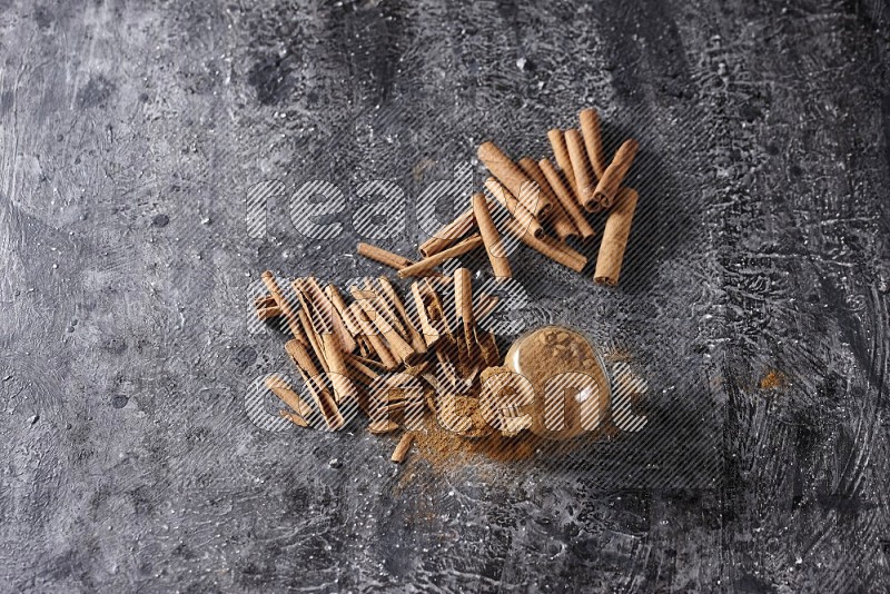 Herbal glass jar full cinnamon powder flipped and a metal spoon full of powder surrounded by cinnamon sticks on textured black background in different angles