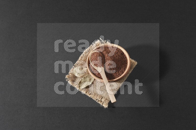 A wooden bowl full of garden cress seeds with wooden spoon full of the seeds on it on burlap fabric on a black flooring