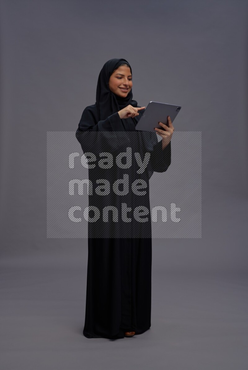 Saudi woman wearing Abaya standing working on tablet on gray background