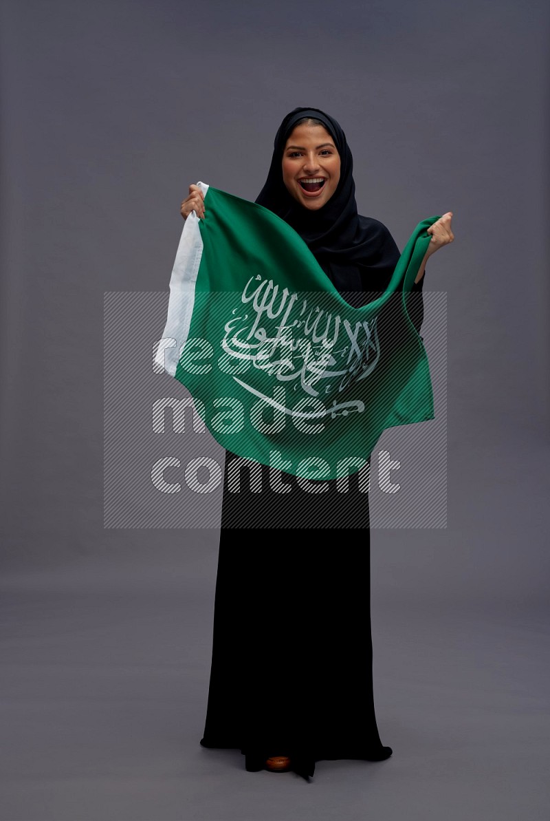 Saudi woman wearing Abaya standing holding Saudi flag on gray background