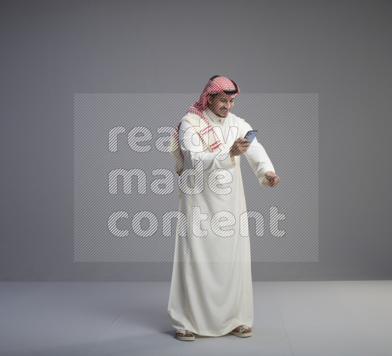 A  Saudi man standing wearing thob and red shomag texting on phone on gray background