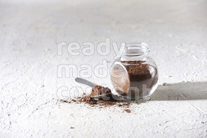 A glass spice jar and a metal spoon full of cloves powder on textured white flooring