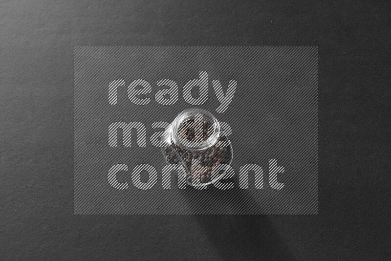 A glass spice jar full of black peppers on black flooring