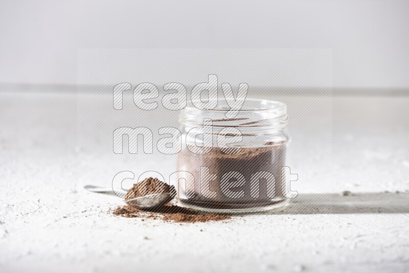 A glass jar full of cloves powder with a metal spoon on a textured white flooring