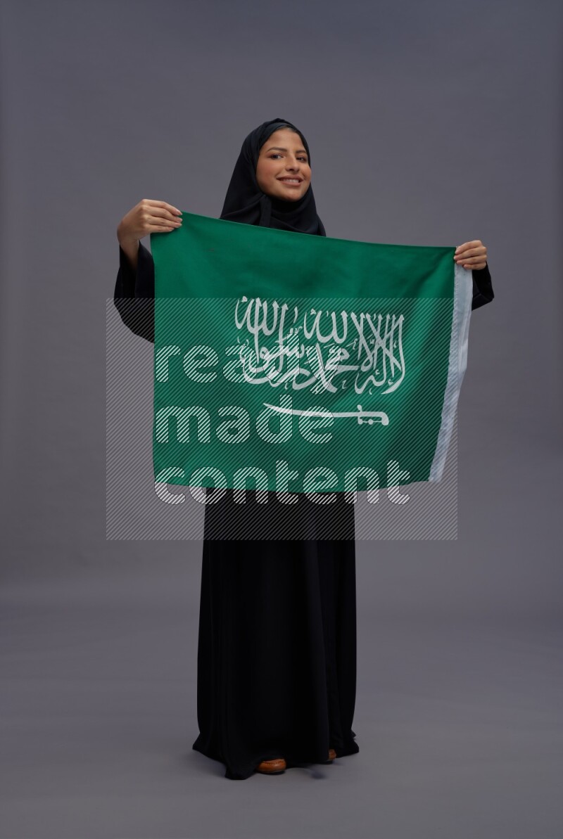 Saudi woman wearing Abaya standing holding Saudi flag on gray background