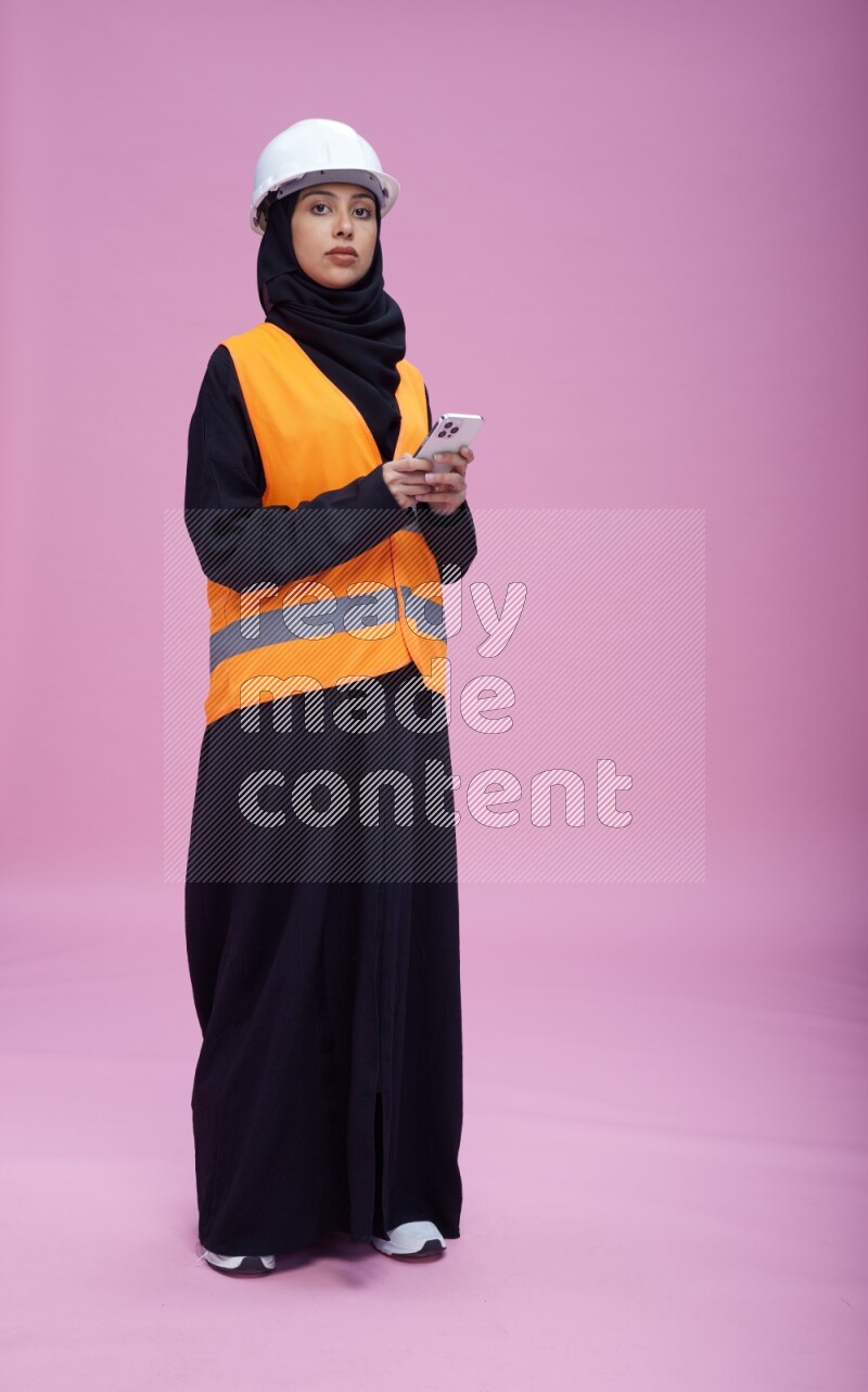 Saudi woman wearing Abaya with engineer vest and helmet standing texting on phone on pink background
