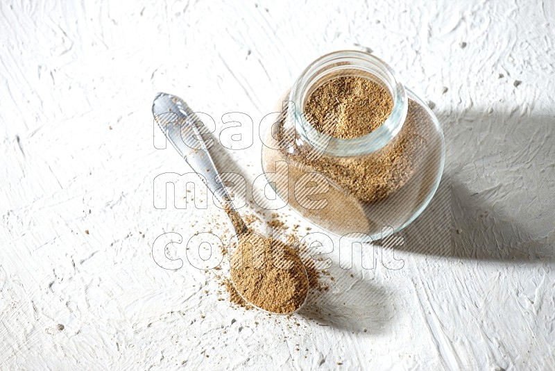 A glass spice jar and metal spoon full of cumin powder on textured white flooring