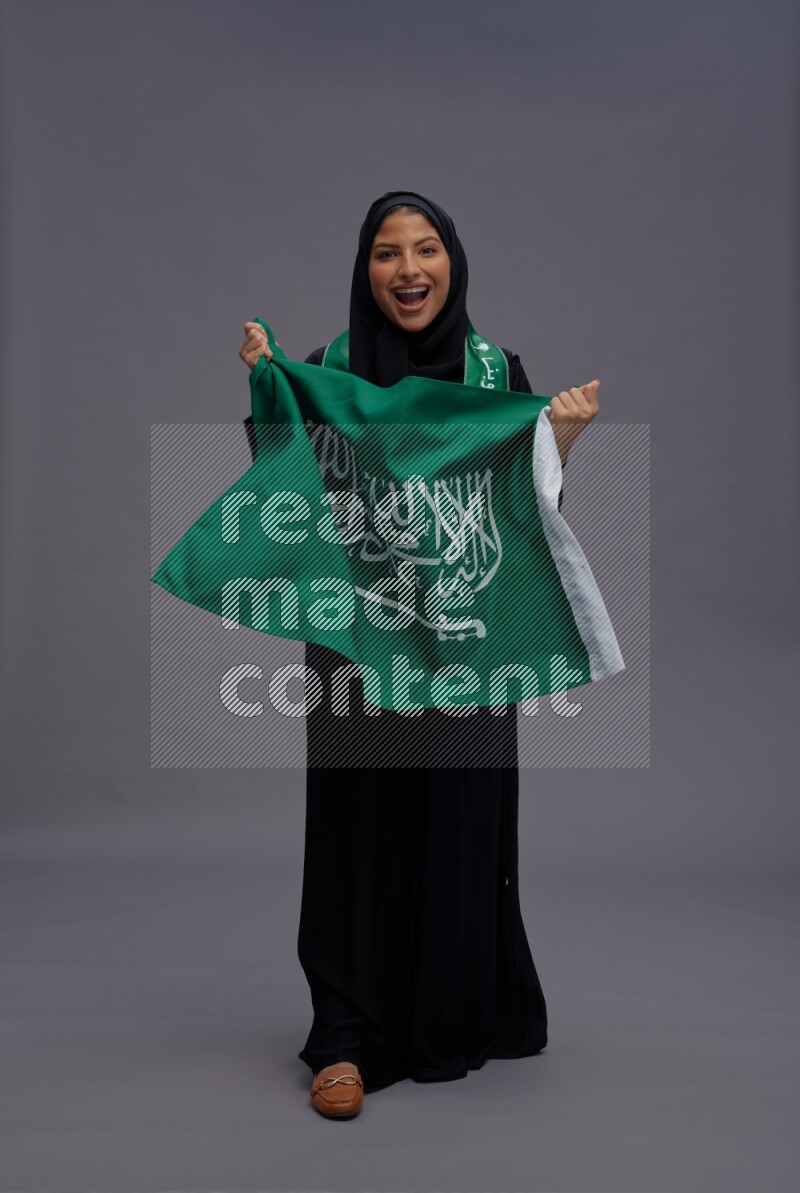 Saudi woman wearing Abaya standing holding Saudi flag on gray background