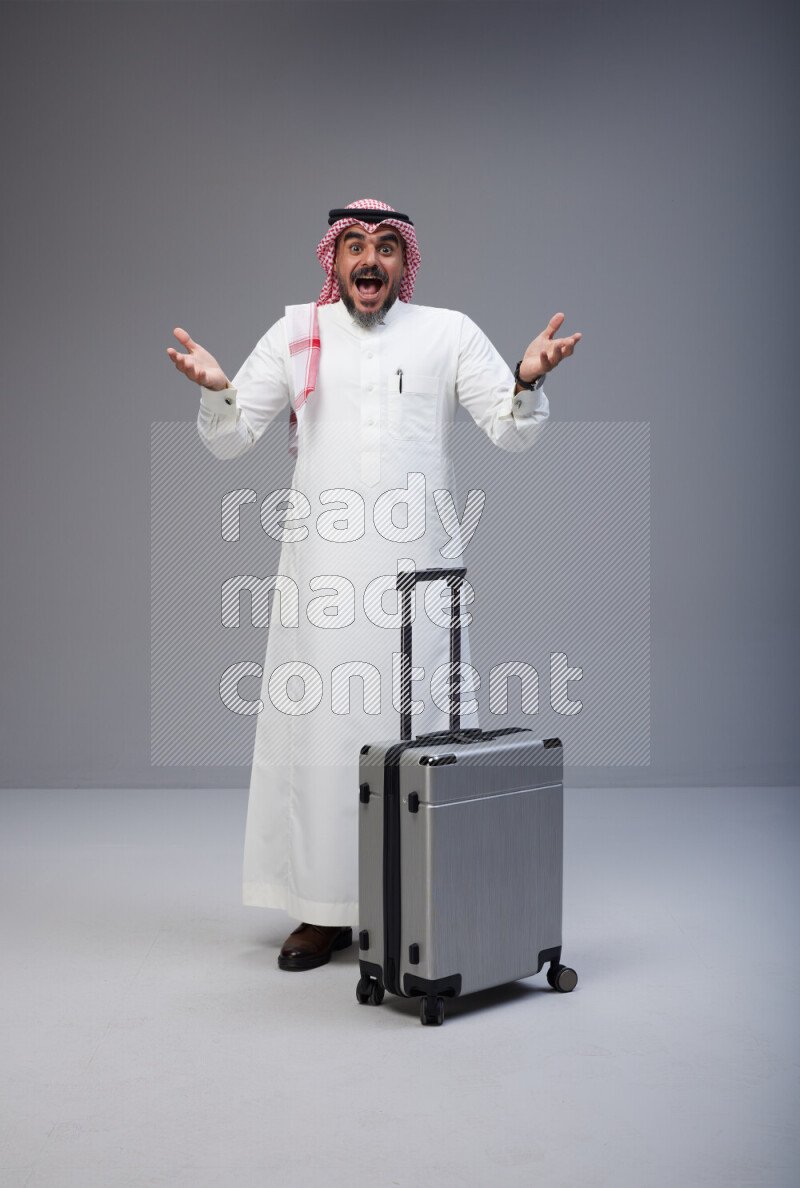 Saudi man wearing Thob and red Shomag standing holding Travel bag on Gray background