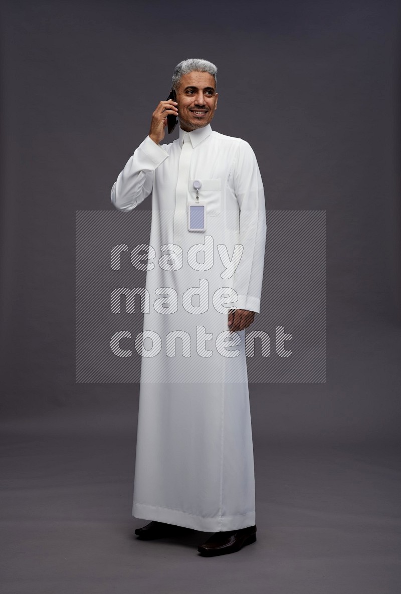 Saudi man wearing thob with pocket employee badge standing talking on phone on gray background