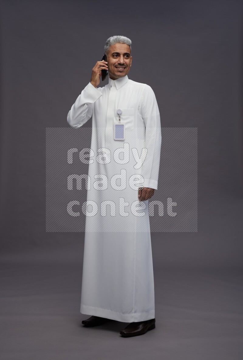Saudi man wearing thob with pocket employee badge standing talking on phone on gray background