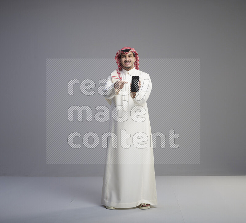 A Saudi man standing wearing thob and red shomag showing phone to camera on gray background