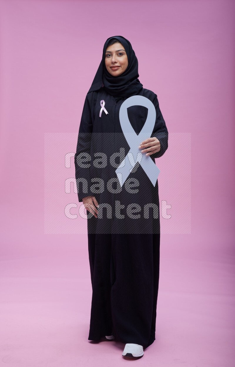 Saudi woman wearing pink ribbon on abaya standing holding awareness ribbon on plain pink background