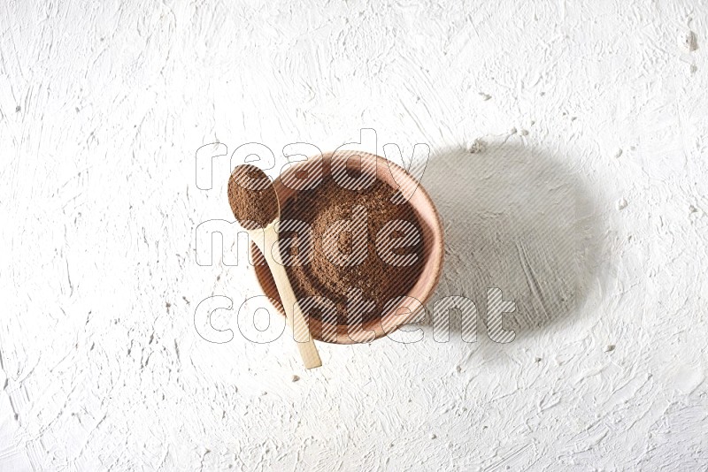 A wooden bowl and a wooden spoon full of cloves powder on a textured white flooring