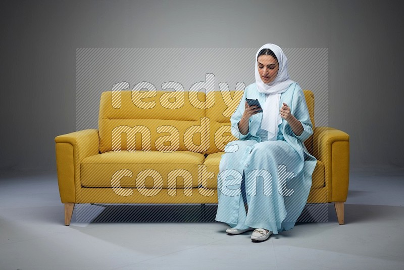 A Saudi woman wearing a light blue Abaya and white head scarf sitting on a yellow sofa and using her phone eye level on a grey background