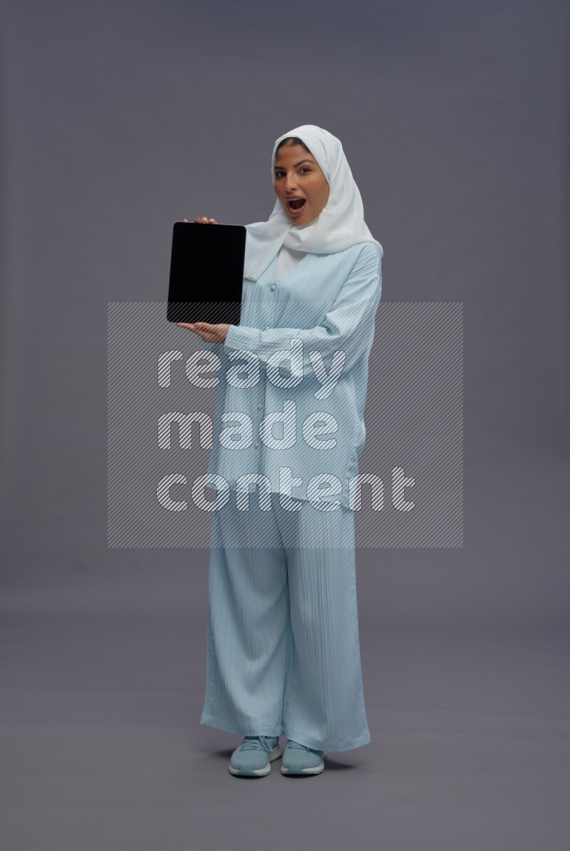 Saudi woman wearing hijab clothes standing showing tablet to camera on gray background