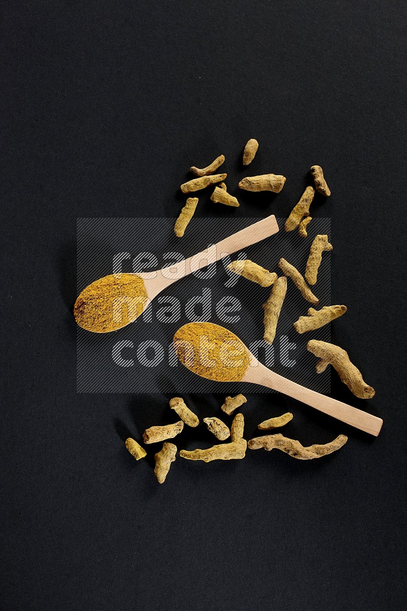 2 wooden spoons full of turmeric powder with dried turmeric fingers on black flooring