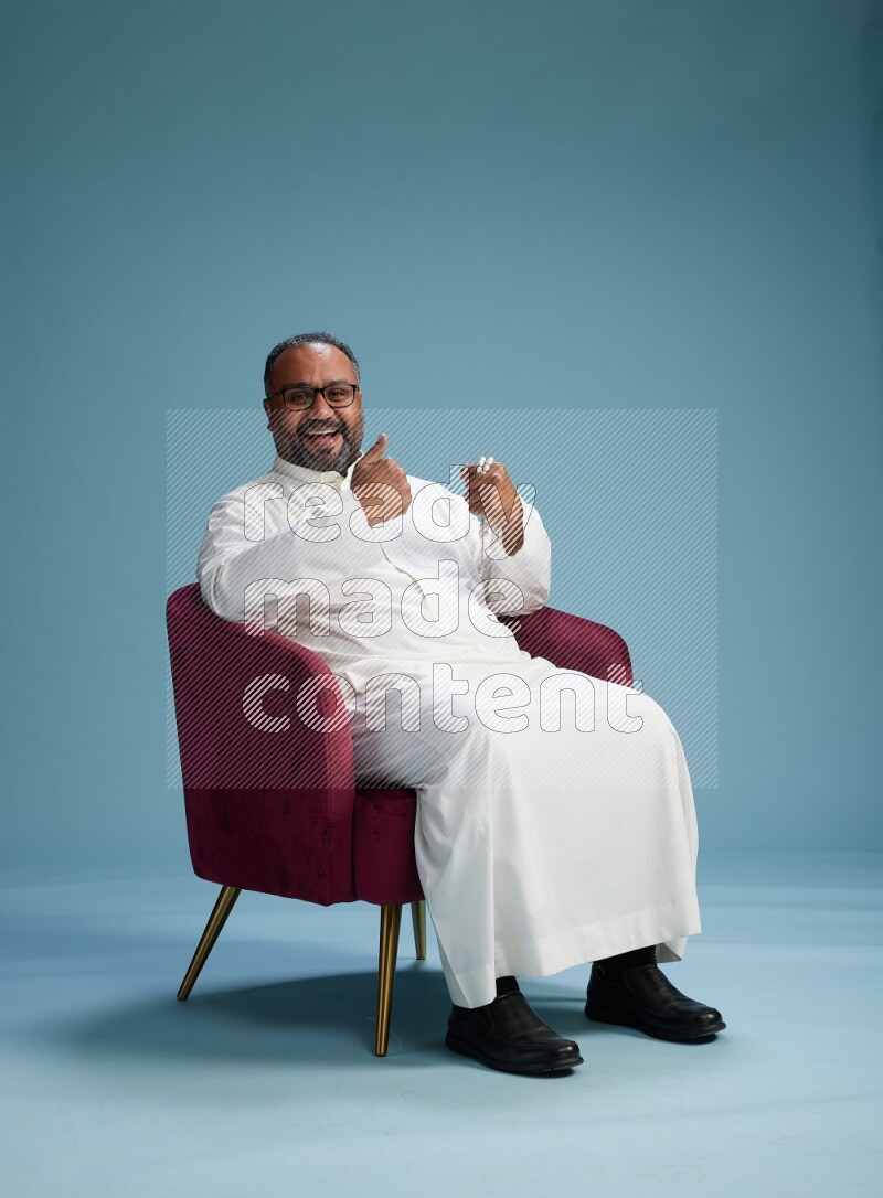Saudi Man without shimag sitting on chair Interacting with the camera on blue background